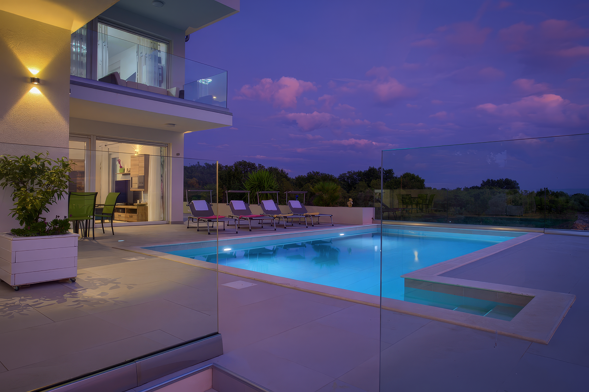 Modern villa in Croatia with a pool illuminated at night, surrounded by lounge chairs and a garden with a view of the natural landscape.